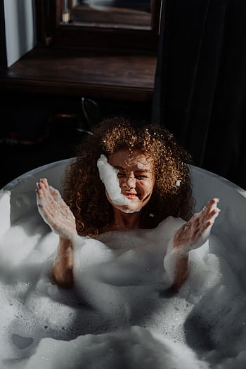 A smiling young woman with curly hair enjoys a relaxing bubble bath in a cozy bathroom setting. HD phone wallpaper
