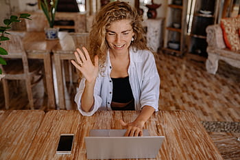 Smiling woman waves during a video call on her laptop in a comfortable indoor setting. HD wallpaper