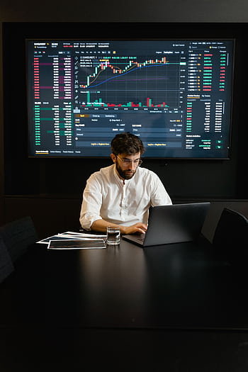 A focused man analyzing stock market trends on a laptop in an office setting with charts displayed. HD phone wallpaper