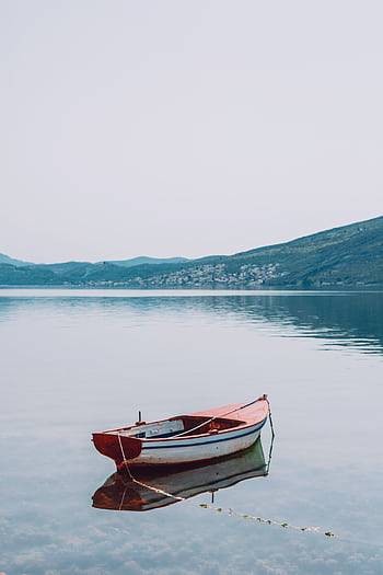 A solitary red boat floats on a calm lake against a backdrop of distant hills. HD phone wallpaper