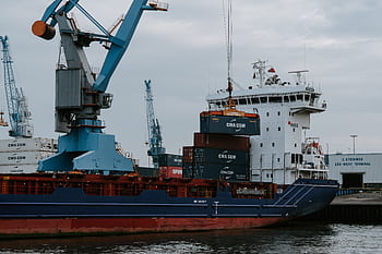 A cargo ship loaded with shipping containers at an industrial harbor with cranes. HD wallpaper