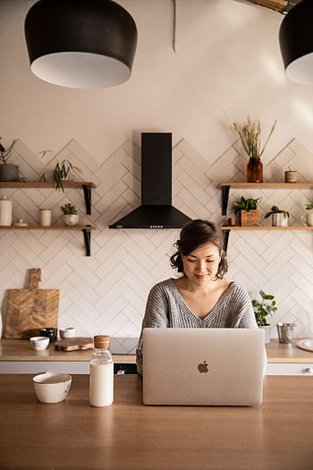 A young woman uses a laptop in a stylish kitchen, embodying remote work lifestyle. HD phone wallpaper