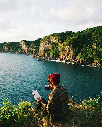 Person enjoys a peaceful moment, reading by the ocean on a cliffside, surrounded by nature. HD phone wallpaper