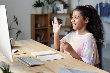 A happy girl waves during an online class at home, sitting at a desk with a computer. HD wallpaper