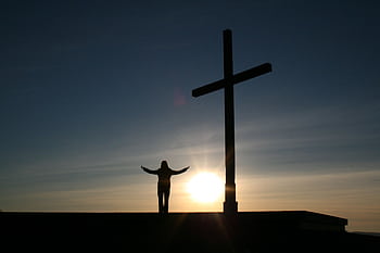 Silhouette of a person with open arms beside a cross at sunset, symbolizing faith and spirituality. HD wallpaper