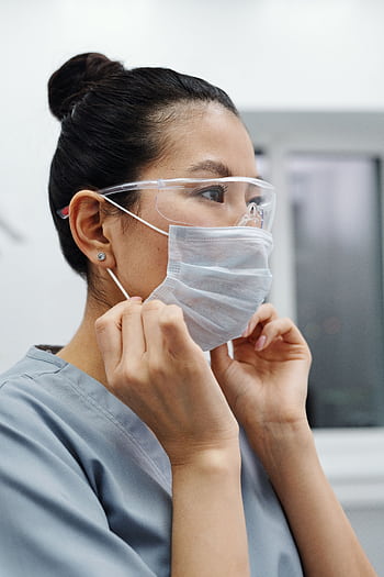 Asian female healthcare worker putting on a protective mask indoors, emphasizing health and safety. HD phone wallpaper