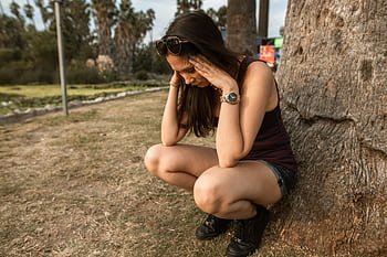 A woman kneels by a tree holding her head, expressing stress or anxiety in an outdoor park. HD wallpaper