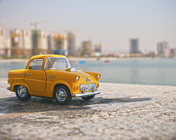 A bright yellow toy car set against a blurred city skyline beside a water body. HD wallpaper