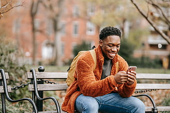 A cheerful young man in warm clothes smiling while using his on a bench in a city park. HD wallpaper