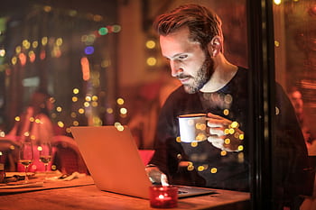 A young man with a beard is working on a laptop at a cozy cafe with warm lighting, holding a coffee cup. HD wallpaper