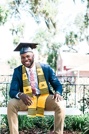 Smiling graduate sitting outside wearing a cap and gown with stole. HD phone wallpaper