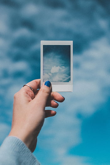 A hand with painted nails holds a Polaroid of clouds against a vibrant blue sky background. HD phone wallpaper