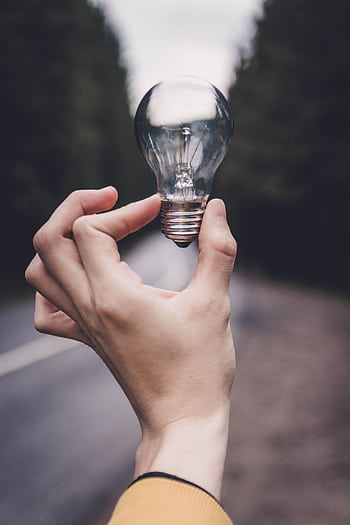 Close-up of a hand holding a lightbulb against a blurred outdoor road background. HD phone wallpaper