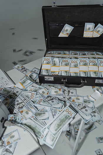 A briefcase overflowing with neatly stacked US dollar bills surrounded by scattered currency on a table. HD phone wallpaper