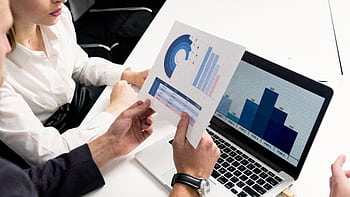 Business professionals discussing data charts and graphs in a modern office setting. HD wallpaper