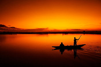 A serene silhouette of fishermen navigating a boat across a calm lake during a vivid sunset. HD wallpaper