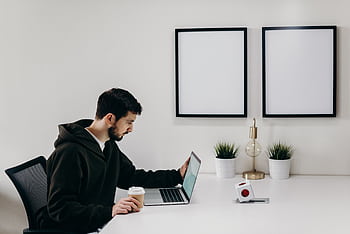 Young man in a hoodie works on a laptop in a modern, minimalist home office setup. HD wallpaper