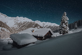 A serene winter night with cabins blanketed in snow, beneath an expansive starry sky and snow-capped mountains. HD wallpaper