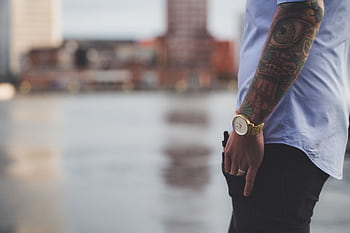 Close-up of a tattooed arm with watch against blurred city backdrop, capturing modern urban style. HD wallpaper
