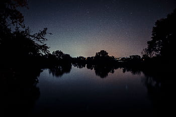 Peaceful lakeside view with starry night sky reflecting on calm waters, creating a serene atmosphere. HD wallpaper