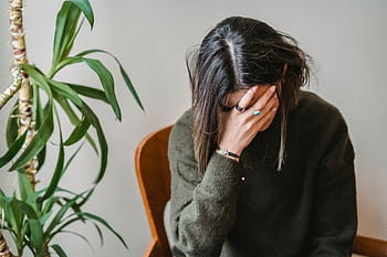 A young woman sits indoors covering her face with her hand, conveying emotions of stress and contemplation. HD wallpaper