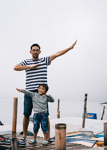 Father and son posing playfully on a wooden dock by the water, enjoying a day outdoors. HD phone wallpaper