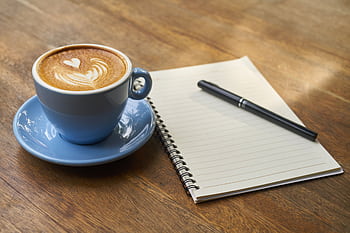 A calming scene of a latte with art in a blue cup beside a notebook and pen on a wooden table. HD wallpaper