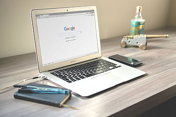 A neat workspace featuring a laptop displaying Google search, a smart, and a notebook on a wooden desk. HD wallpaper