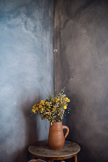 A rustic display of dried yellow flowers in a terracotta vase on a wooden table against a textured wall. HD phone wallpaper
