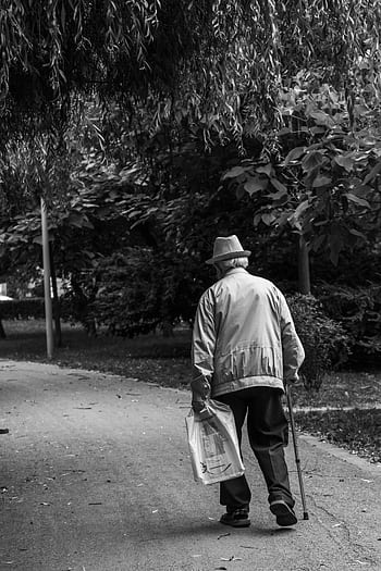 Elderly man in a hat walking on a park path with a cane, captured in monochrome. HD phone wallpaper