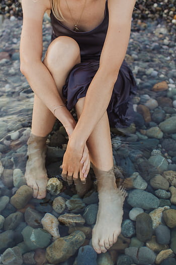 A woman sits with her feet in rocky beach waters, enjoying a serene moment. HD phone wallpaper