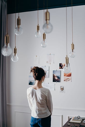 A woman stands inside, viewing an inspiring mood board, under hanging lights. HD phone wallpaper