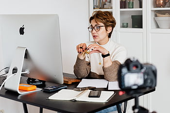 A professional woman engaged in a virtual meeting setup at her home desk, using a computer and camera. HD wallpaper