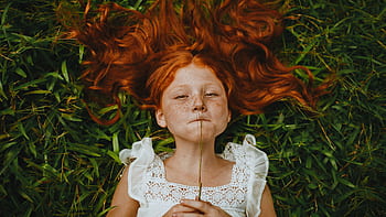 A young redhead girl lies in the grass holding a blade of grass, showcasing her freckles and peaceful expression. HD wallpaper