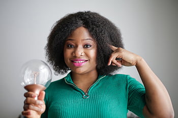 Smiling black woman with afro hair holding a light bulb against a gray background, symbolizing creativity and inspiration. HD wallpaper
