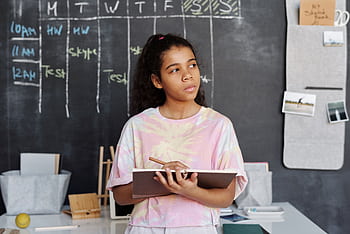 A teenager in a pink shirt ponders while holding a notebook in a classroom setting. HD wallpaper