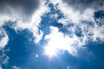 A vivid view of the sun shining through fluffy clouds in a bright blue sky. HD wallpaper
