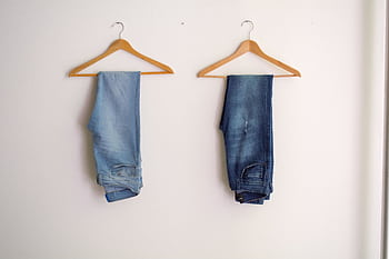 Two pairs of blue denim jeans neatly hanging on wooden hangers against a plain wall. HD wallpaper