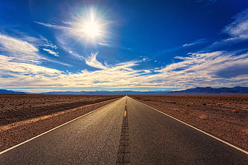 A long, straight road stretches across a dry desert landscape under a bright blue sky and shining sun. HD wallpaper