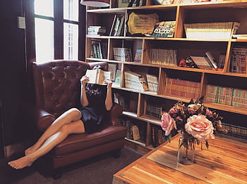 Woman relaxing with a book in a cozy library armchair, surrounded by wooden bookshelves and flowers. HD wallpaper