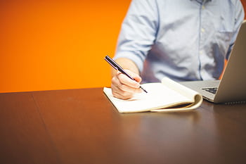 A person writing notes at a workspace with a laptop and vibrant orange background. HD wallpaper