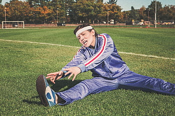 A woman in a tracksuit stretching on a sports field, surrounded by greenery. HD wallpaper