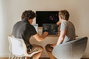 Two male developers working on laptops indoors, discussing code. HD wallpaper