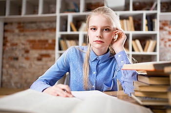 A young woman with blond hair studying at a table piled with books. HD wallpaper