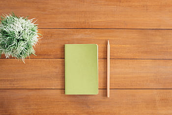 Flat lay of green notebook and pencil on wooden desk with plant. Perfect for creativity and organization themes. HD wallpaper