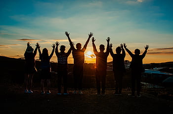 A diverse group of friends raises their arms in celebration against a vibrant sunset backdrop. HD wallpaper