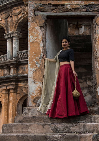 A woman in a vibrant red skirt and black blouse stands gracefully on rustic stone stairs in an ancient Indian setting. HD phone wallpaper