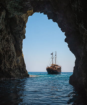 A stunning view of a ship sailing through a rocky sea cave in Rethymno, Greece. HD phone wallpaper