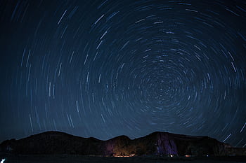 A mesmerizing night sky showcasing rotating star trails over a mountainous landscape. HD wallpaper