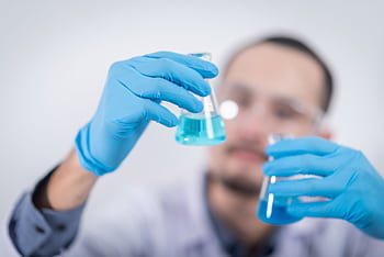 Scientist in a laboratory conducting an experiment, holding blue liquid in flasks with protective gloves. HD wallpaper
